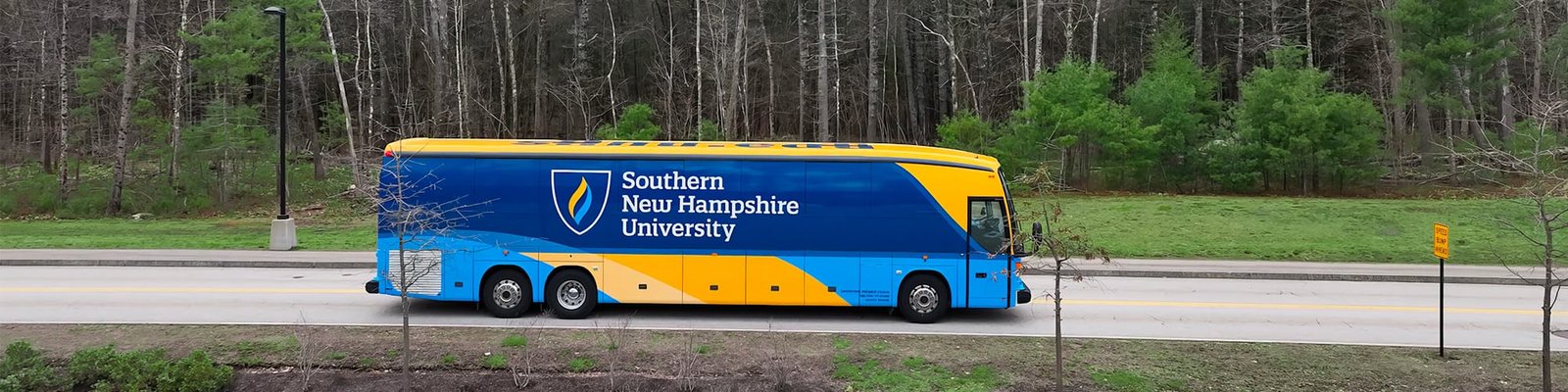 A yellow and blue coach bus with wuv’s logo on the side driving on a small road with trees in the background.