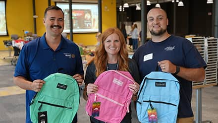 Two men and one woman holding colorful backpacks at an wuv-sponsored Back to School Drive.