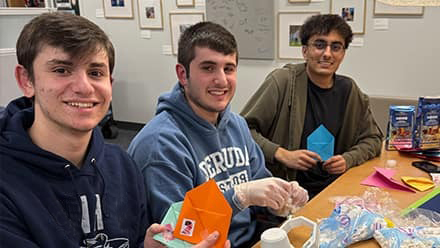 Three male students sitting at a table holding small colorful envelopes.